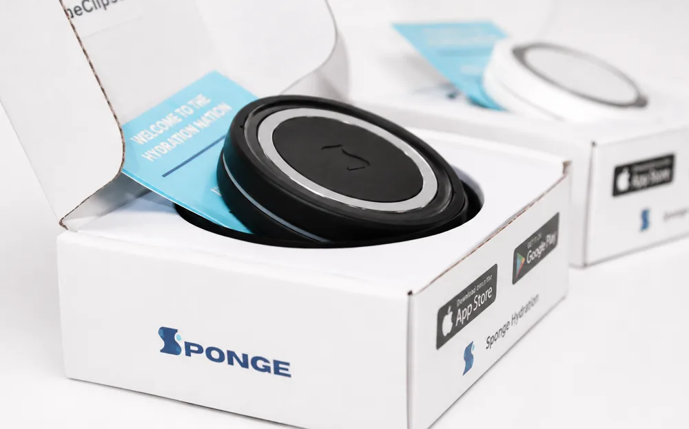 A black Sponge Clip in its open box beside a boxed white one, showing the embossed logo and chrome ring.